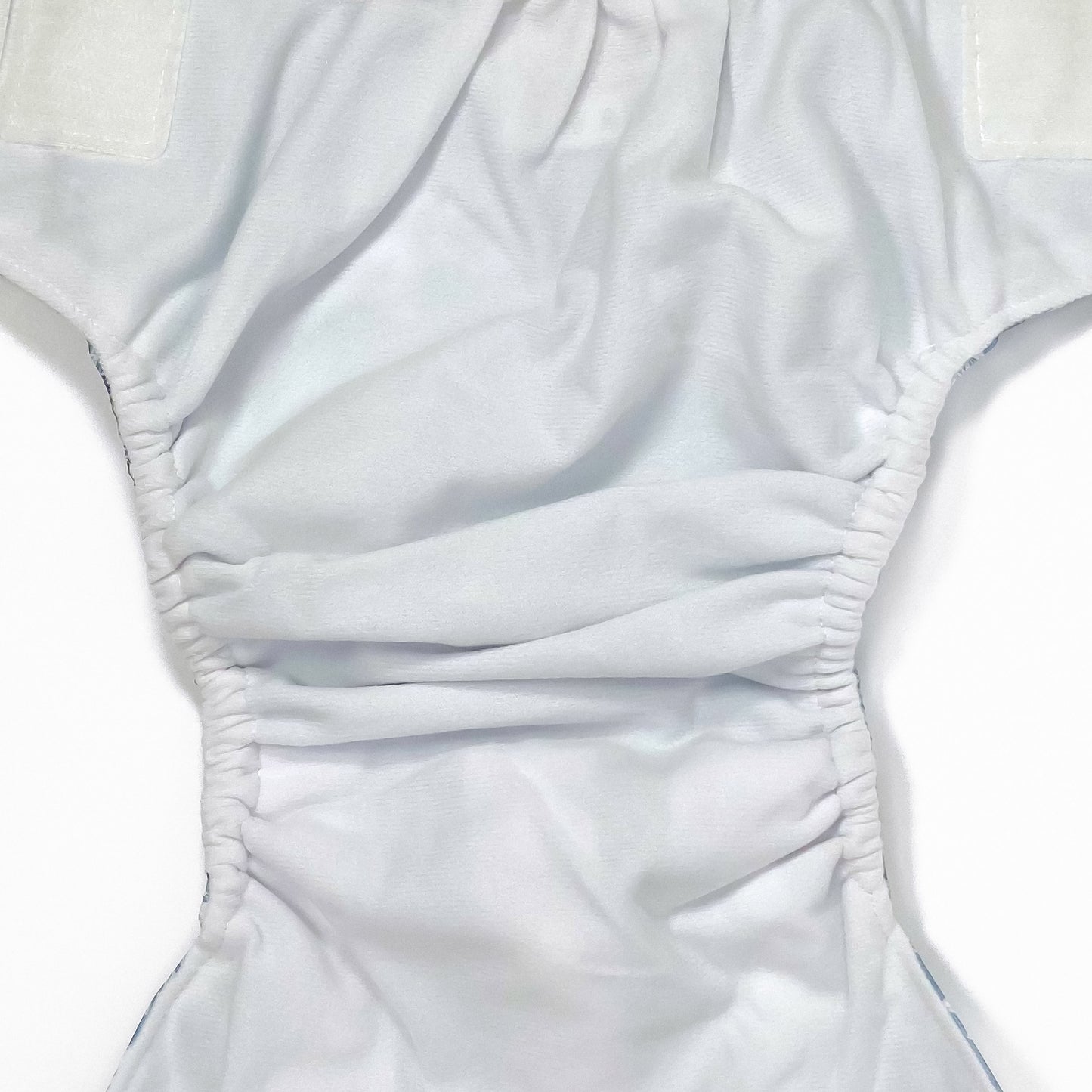 An adjustable reusable nappy for babies and toddlers, featuring a blue fern design, with images of white fern leaves on a blue background. Image shows a zoomed in view of the nappy fabric lining.