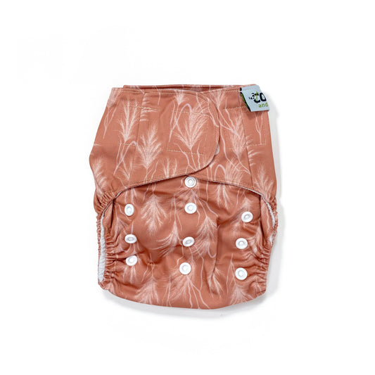 An adjustable reusable nappy for babies and toddlers, featuring a sunset dune design, with images of grasses on a reddish-brown background. View shows the front of the nappy, with fastenings closed.