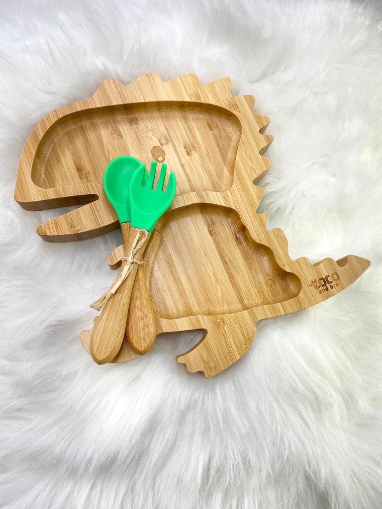 A bamboo children’s plate, with multiple food sections, in a dinosaur design. The bamboo plate features a green silicone suction ring at the back, and includes bamboo fork and spoon utensils in matching colours. Front view.