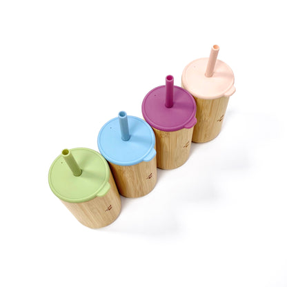 A collection of children’s bamboo drinking cups, with colourful silicone lids and matching straws. View shows the cups from above, arranged in a line displaying the silicone lids and straws.