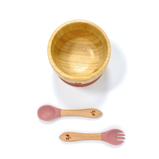 A children’s bamboo bowl with a blush pink silicone suction ring, and matching blush pink silicone and bamboo cutlery.