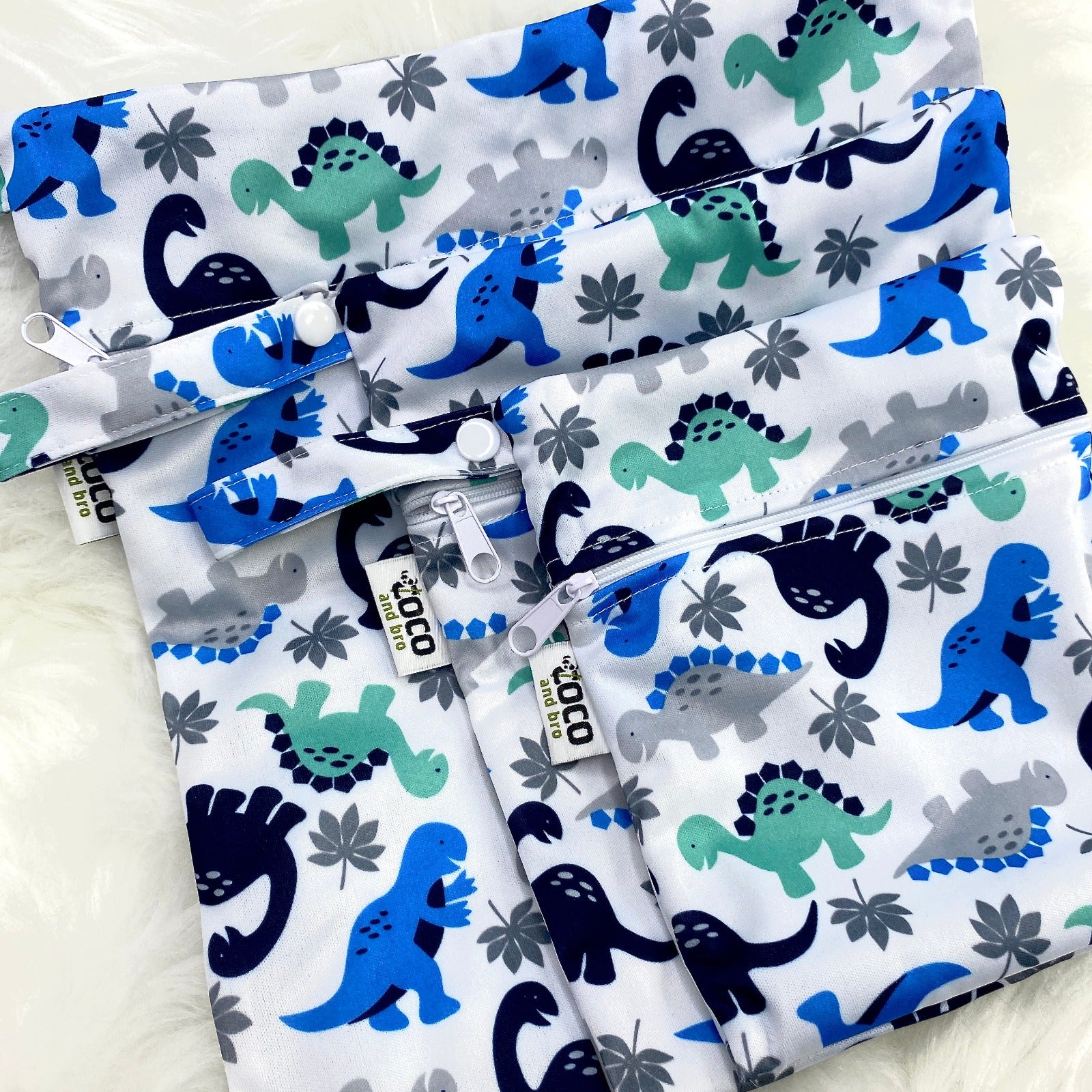 A set of three waterproof bags in a green and blue dinosaur design on a white background, made from bamboo and in three different sizes. Each bag has a zipper closure.