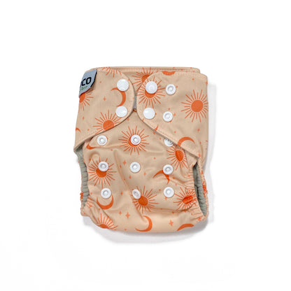 An adjustable reusable nappy for babies and toddlers, featuring a peach cosmos design, with images of moons, suns and stars on a peach background.  View shows the front of the nappy, with fastenings closed.