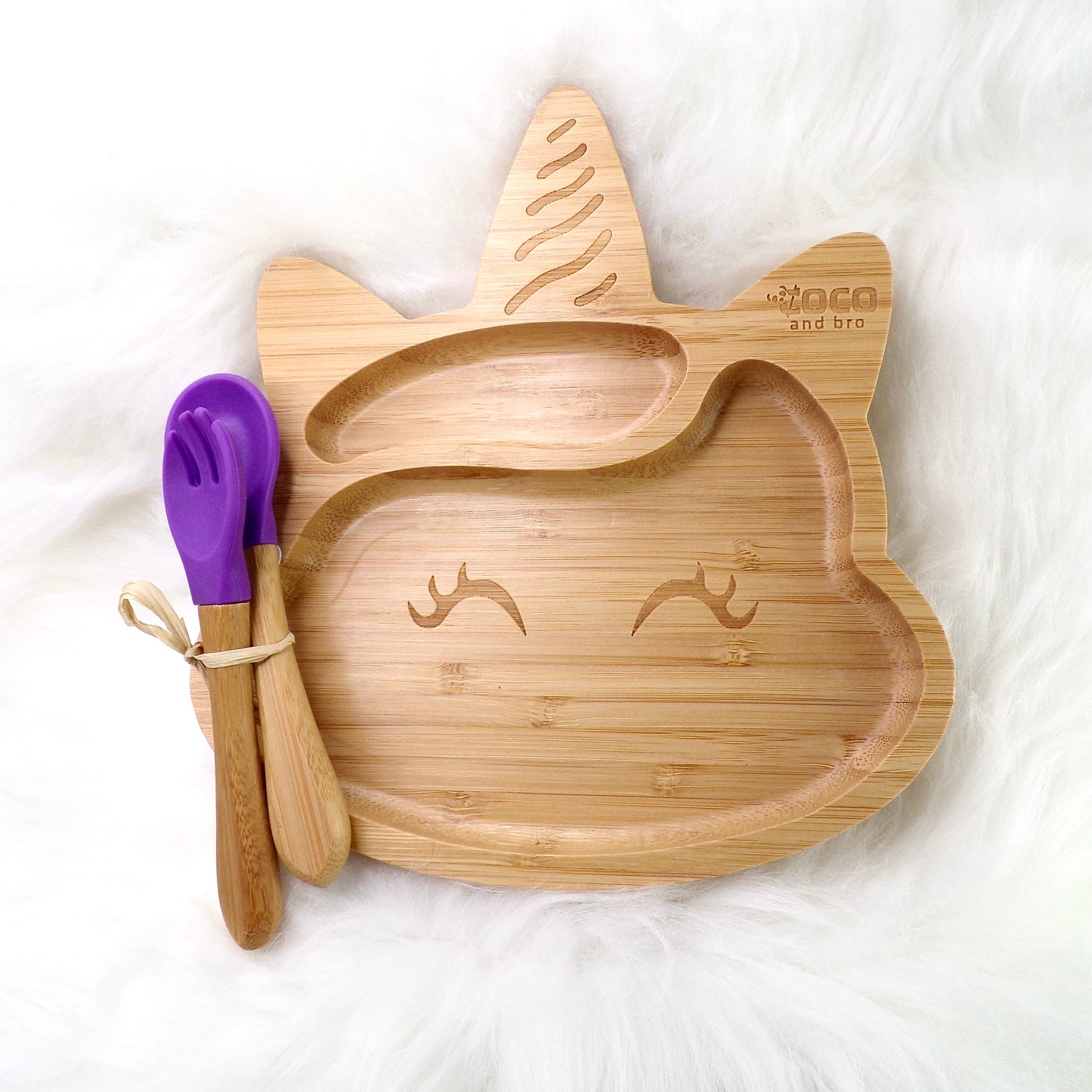 A bamboo children’s plate, with multiple food sections, in a unicorn design. The bamboo plate features a purple silicone suction ring at the back, and includes bamboo fork and spoon utensils in matching colours. Front view.