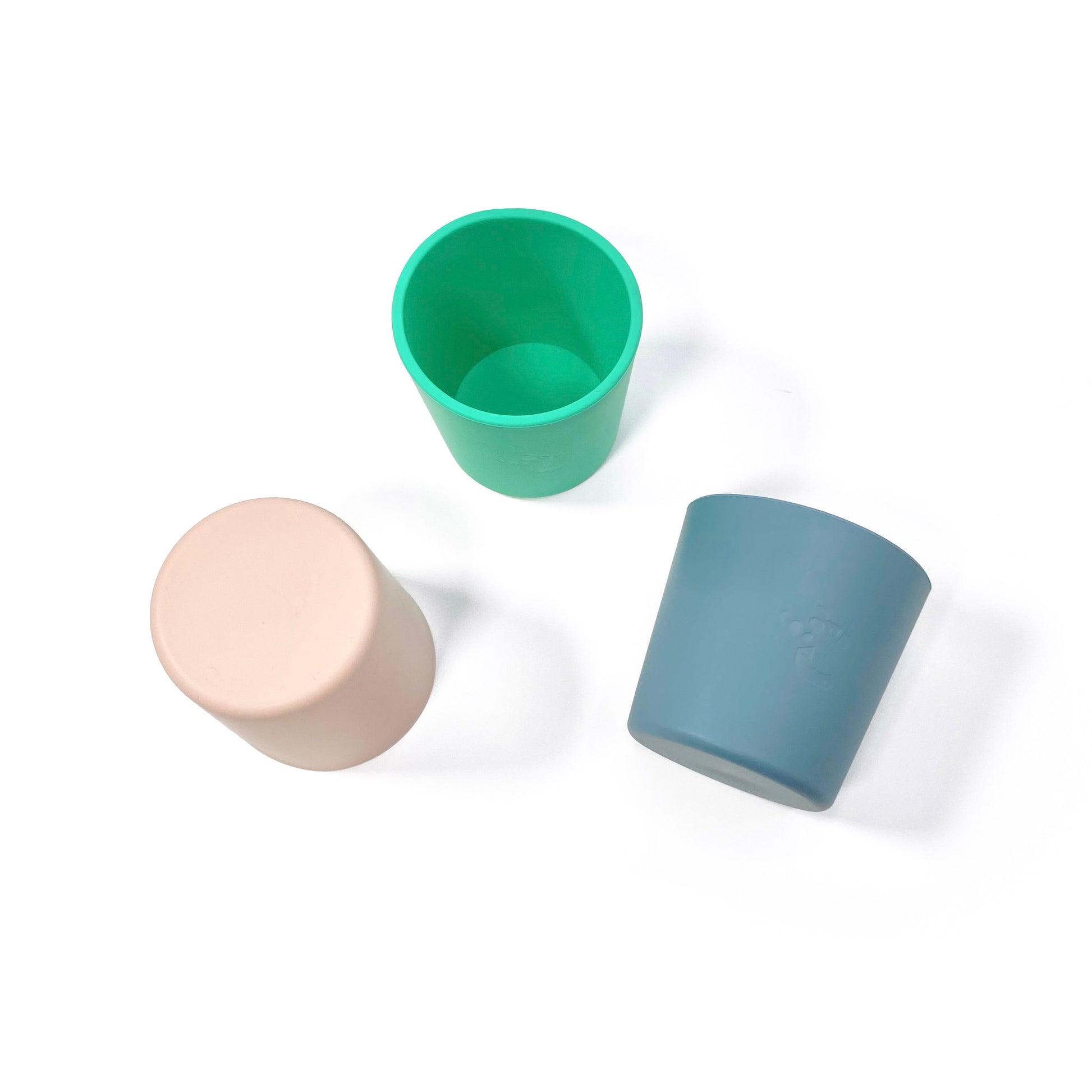 A collection of silicone children’s cups, available in peachy pink, ocean blue and seafoam green. The cups are shown in a scattered formation. View from above.