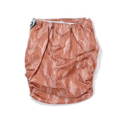 An adjustable reusable nappy for babies and toddlers, featuring a sunset dune design, with images of grasses on a reddish-brown background. View shows the back of the nappy.