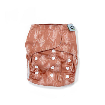 An adjustable reusable nappy for babies and toddlers, featuring a sunset dune design, with images of grasses on a reddish-brown background. View shows the front of the nappy.