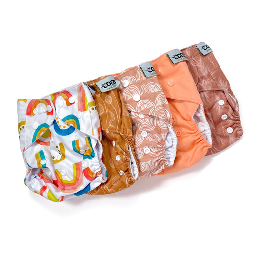 A set of five reusable nappies, made from 100% recycled polyester. The included nappies are: Bright Rainbow, Copper Bloom, Peach Rainbow, Cute Coral and Sunset Dune. View shows all five nappies front facing, in a diagonal line.
