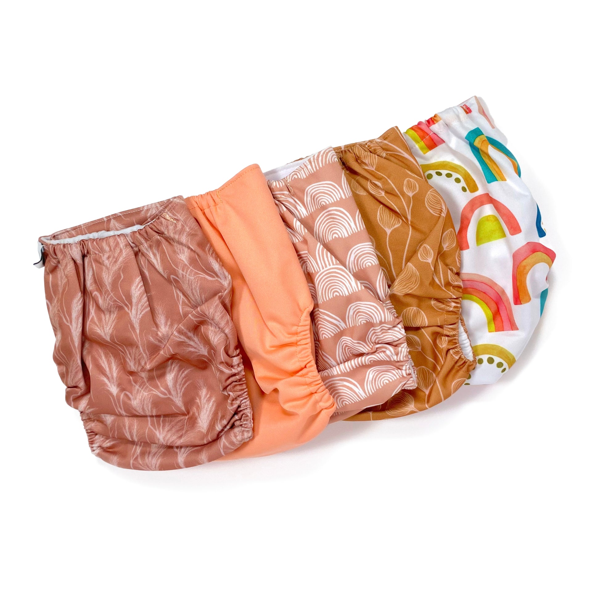 A set of five reusable nappies, made from 100% recycled polyester. The included nappies are: Bright Rainbow, Copper Bloom, Peach Rainbow, Cute Coral and Sunset Dune. View shows all five nappies back facing, in a diagonal line.