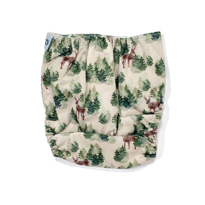 An adjustable reusable nappy for babies and toddlers, featuring a winter forest design with images of deer and pine trees on a cream background. View shows the back of the nappy.