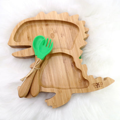 A bamboo children’s plate, with multiple food sections, in a dinosaur design. The bamboo plate features a green silicone suction ring at the back, and includes bamboo fork and spoon utensils in matching colours. Front view.