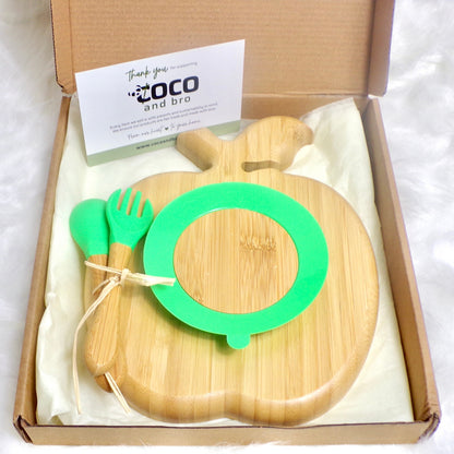 A bamboo children’s plate, with multiple food sections, in an apple design. The bamboo plate features a green silicone suction ring at the back, and includes bamboo fork and spoon utensils in matching colours. Back view. The bamboo plate is presented inside a cardboard box with gift wrapping.
