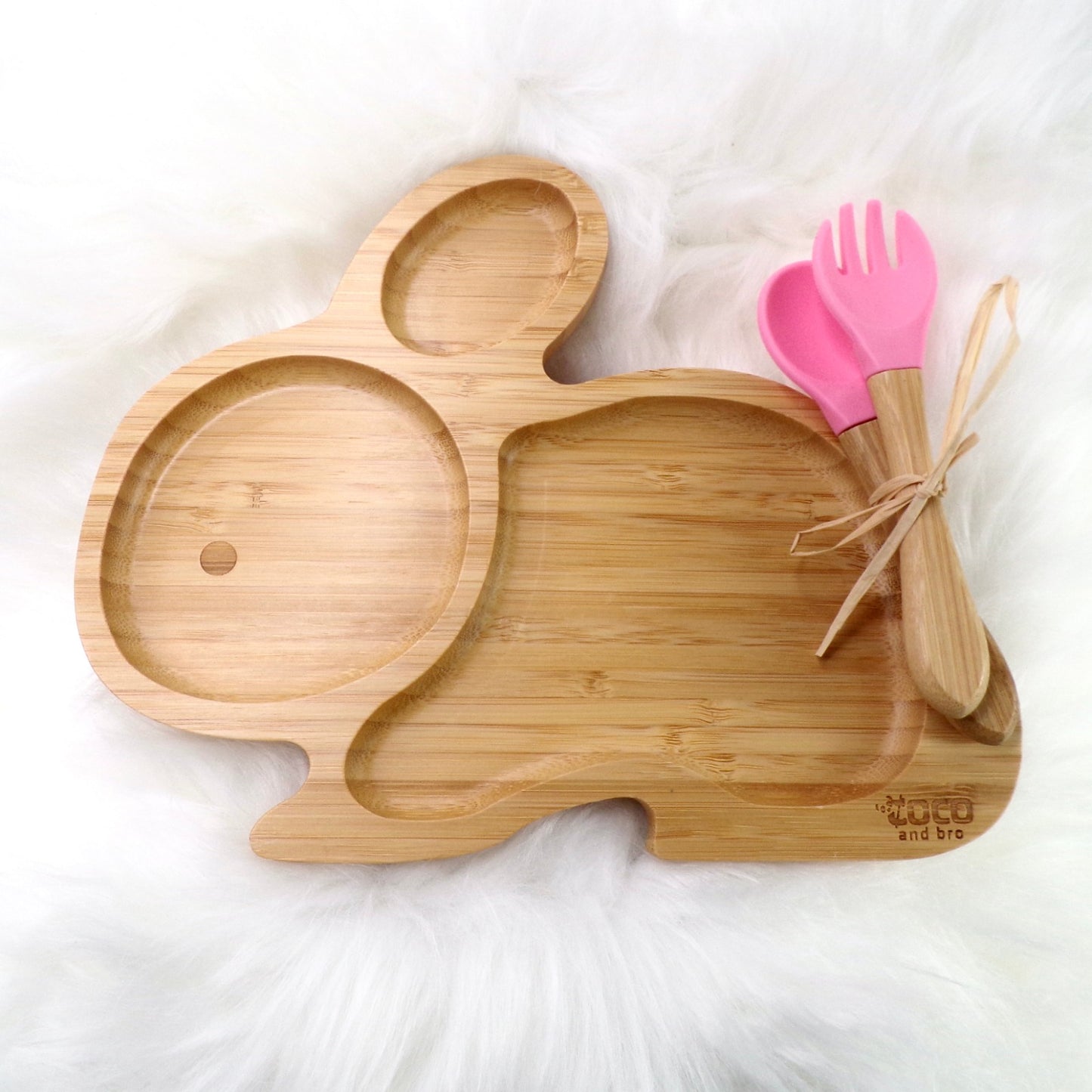 A bamboo children’s plate, with multiple food sections, in a rabbit design. The bamboo plate features a pink silicone suction ring at the back, and includes bamboo fork and spoon utensils in matching colours. Front view.